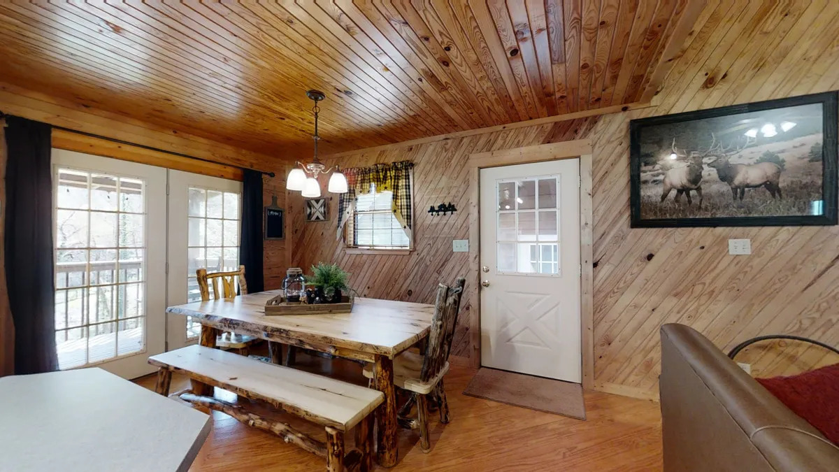 A rustic dining area featuring a large wooden table, soft chandelier lighting, and double doors that open to the outdoors. The setup is ideal for family dinners, game nights, and group celebrations.