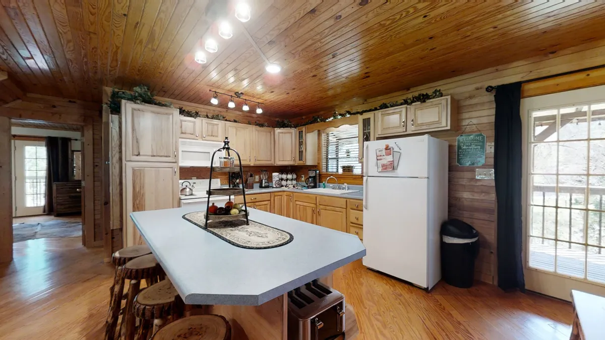A welcoming kitchen with a central island, warm wooden cabinetry, a modern refrigerator, and generous natural light streaming through large windows. The space is set up for cooking and gathering, evoking a sense of comfort and togetherness.