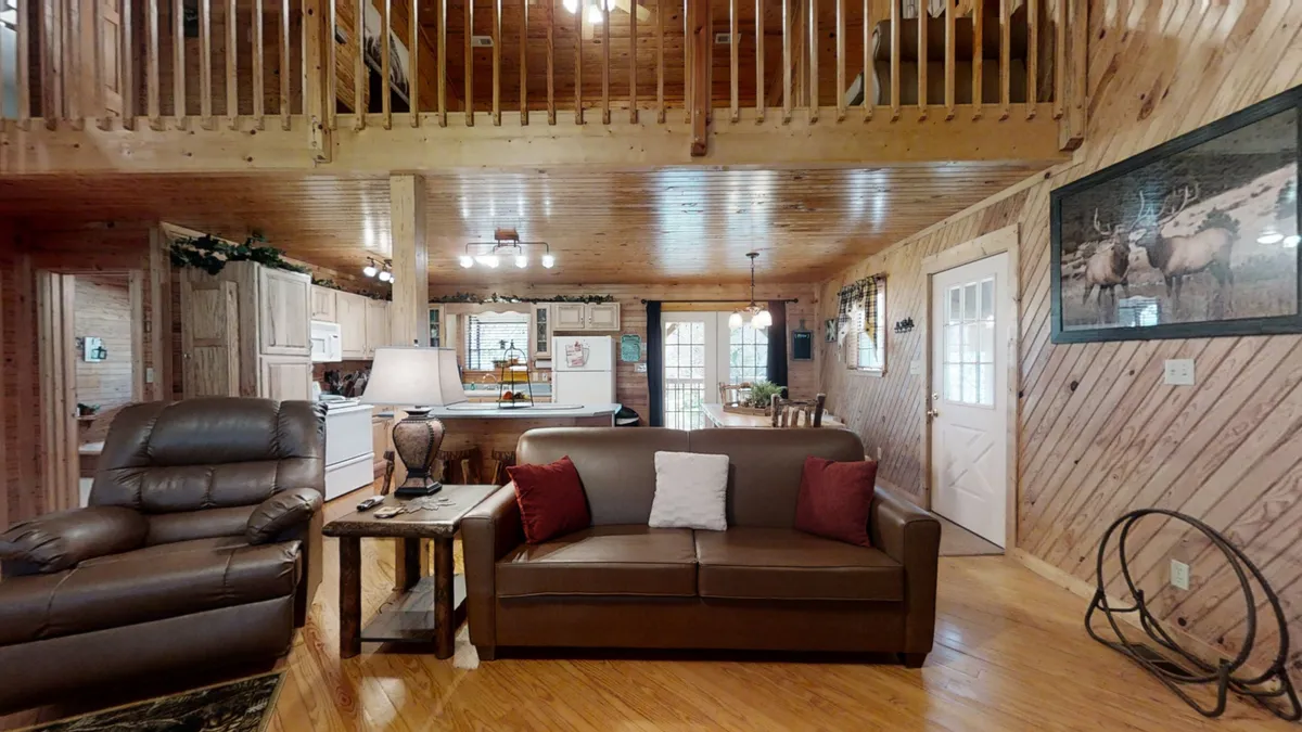 A spacious living area is shown with a plush brown leather sofa, recliner chair, and a wooden coffee table. Large windows bathe the room in natural light, while the open kitchen creates a seamless flow between living and dining areas. The mood is inviting, ideal for relaxing, socializing, or cozy movie nights. The presence of a TV and comfortable seating supports a range of activities.