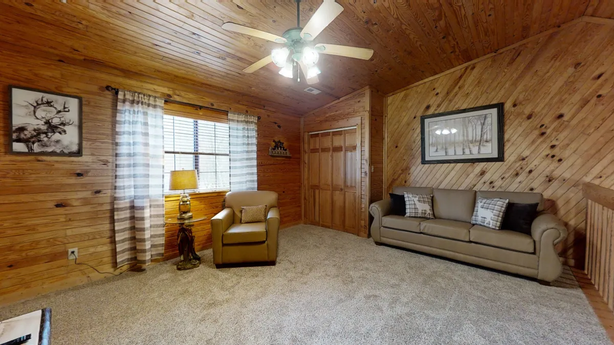 A welcoming loft-style area with wooden walls, a sofa, armchair, lamp, and a window. The arrangement invites relaxation or socializing, with the window providing a view and soft natural illumination.