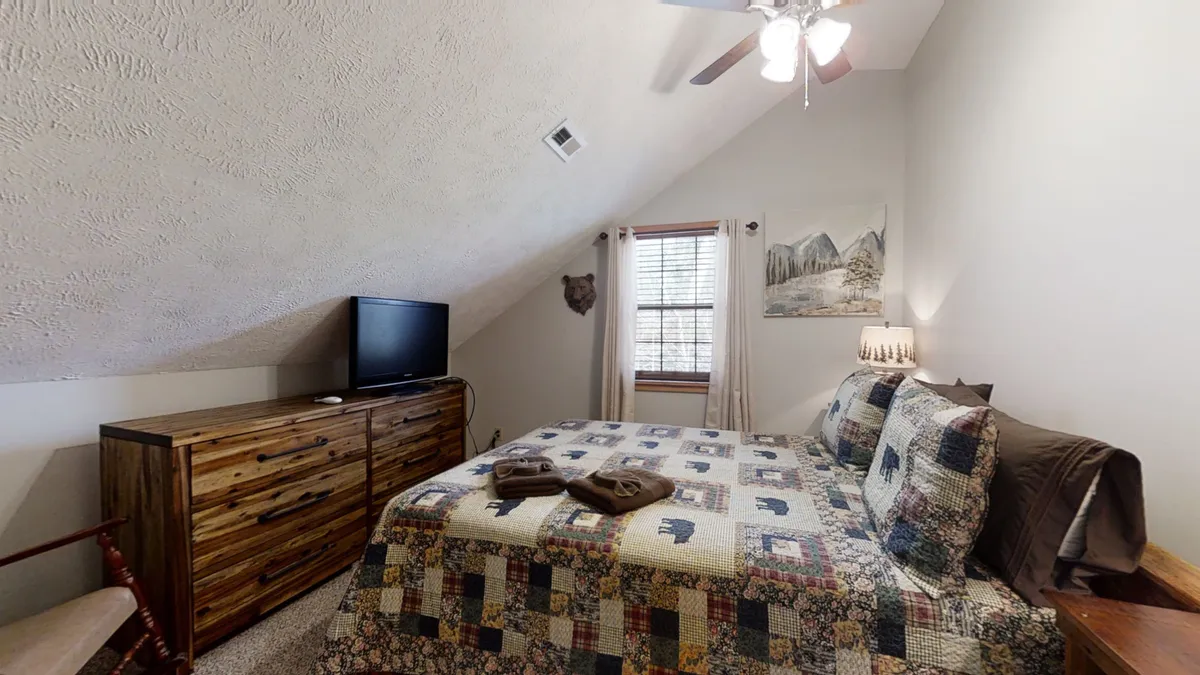 A cozy bedroom featuring a comfortable bed, a dresser, a television, a window with views, and mountain-themed artwork. This space exudes relaxation, offering both entertainment and opportunities to unwind with nature views.