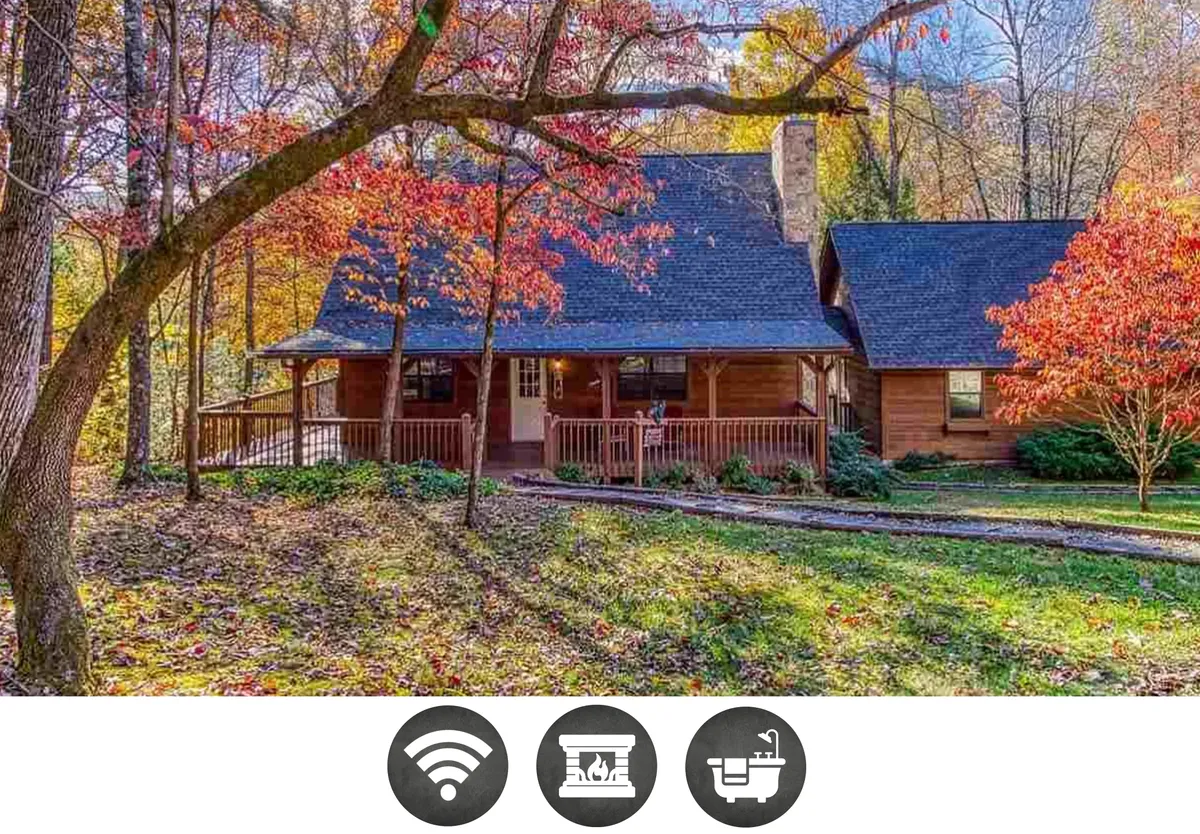 The image showcases the cabin’s inviting wooden porch nestled among vibrant, colorful trees, with a prominent stone chimney adding rustic charm. The setting is tranquil and picturesque, surrounded by nature—perfect for relaxing, enjoying the changing seasons, and gathering with family or friends.