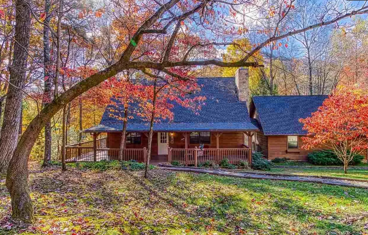 A charming log cabin nestled amidst vibrant autumn foliage, with a welcoming front porch and a sturdy stone chimney. The scene radiates warmth and is clearly designed for families to gather outside, enjoy the natural beauty, and create memories together.