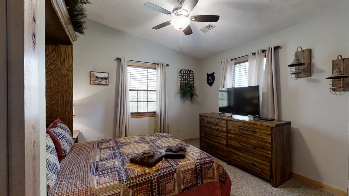 A bright, inviting bedroom with a bed featuring quilted bedding, a wooden dresser, a TV, and a window with curtains. The setup is conducive for relaxing, watching TV, or reading, providing a sense of space and comfort.