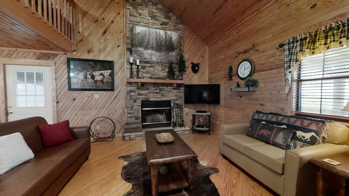 This image centers on a cozy living room with a prominent stone fireplace, a large artwork depicting a forest, a wooden coffee table, a comfortable couch, and an armchair. The space is styled with warm tones and rustic decor, evoking the inviting ambiance of a mountain cabin. The fire flickers in the hearth, and the room feels ready for gatherings or quiet relaxation. Large windows allow in natural light, further enhancing the warm, communal atmosphere.