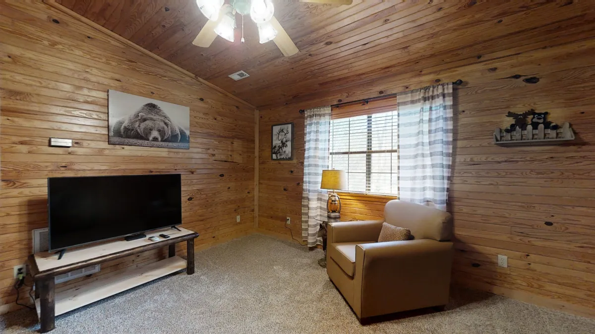 This image depicts a cozy interior featuring a TV on a wooden stand, an armchair, a window with curtains allowing in natural light, and a bear picture on the wall. The space is arranged for relaxation and entertainment, with seating facing the TV and ample light for reading or lounging.