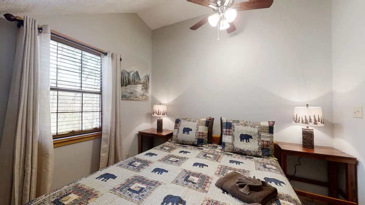 A tranquil bedroom with a bed adorned with a patterned quilt and decorative pillows, flanked by wooden nightstands with warm lamps. Wall art adds a homely touch, and sunlight streams in through a window, creating a serene and restful atmosphere.