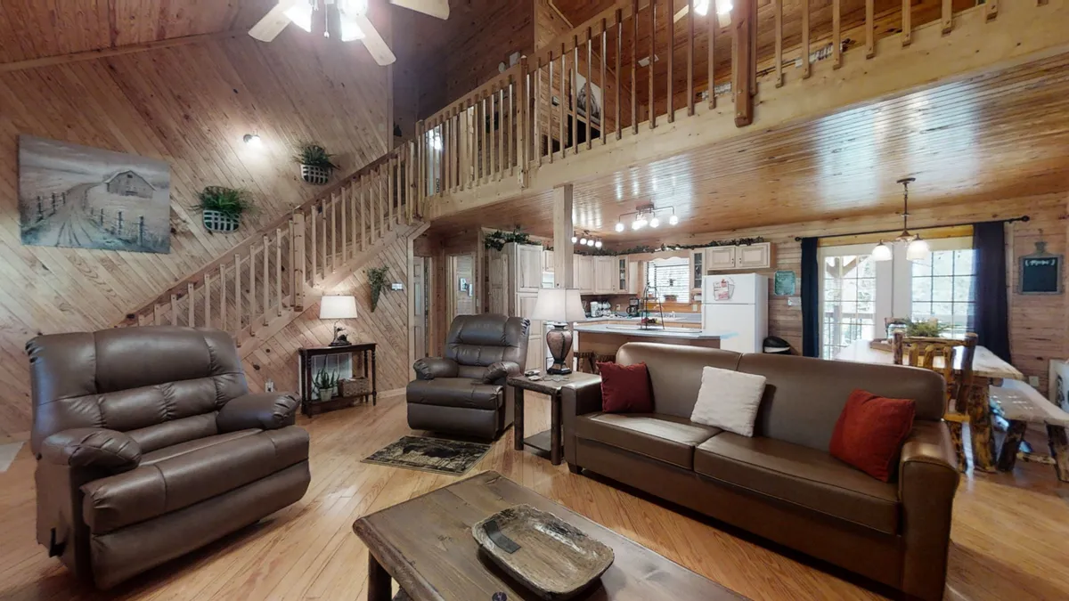 A warm, rustic living room with leather sofas arranged for conversation, a coffee table, and a wooden staircase leading to other parts of the cabin. The open kitchen is visible in the background, reinforcing the home's communal design. The lighting is soft and golden, enhancing the cozy, inviting atmosphere perfect for gatherings or quiet evenings.
